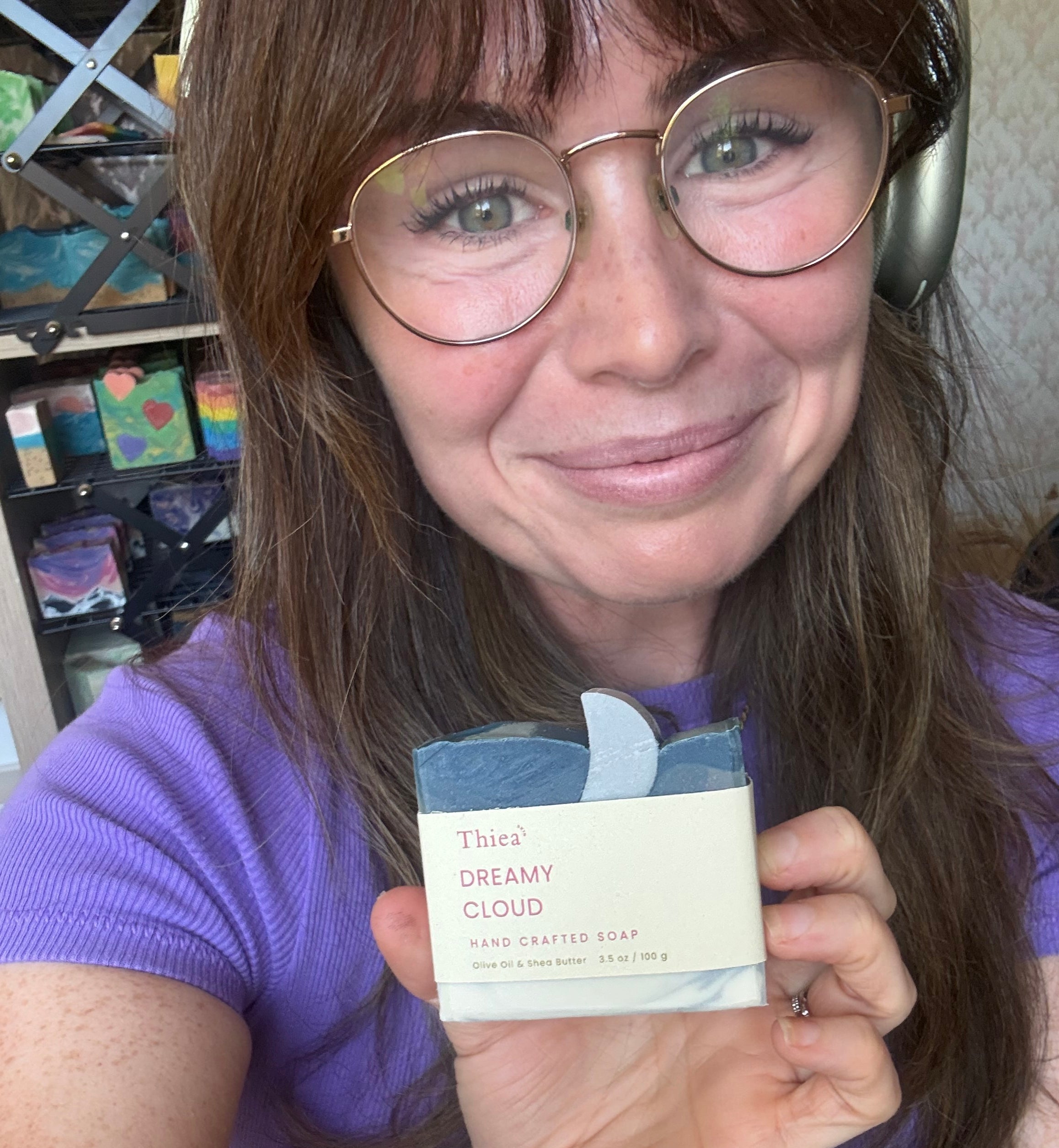 A woman with glasses and a purple shirt holds up a bar of soap labeled 'THAT'S DREAMY CLOUD' next to her face.