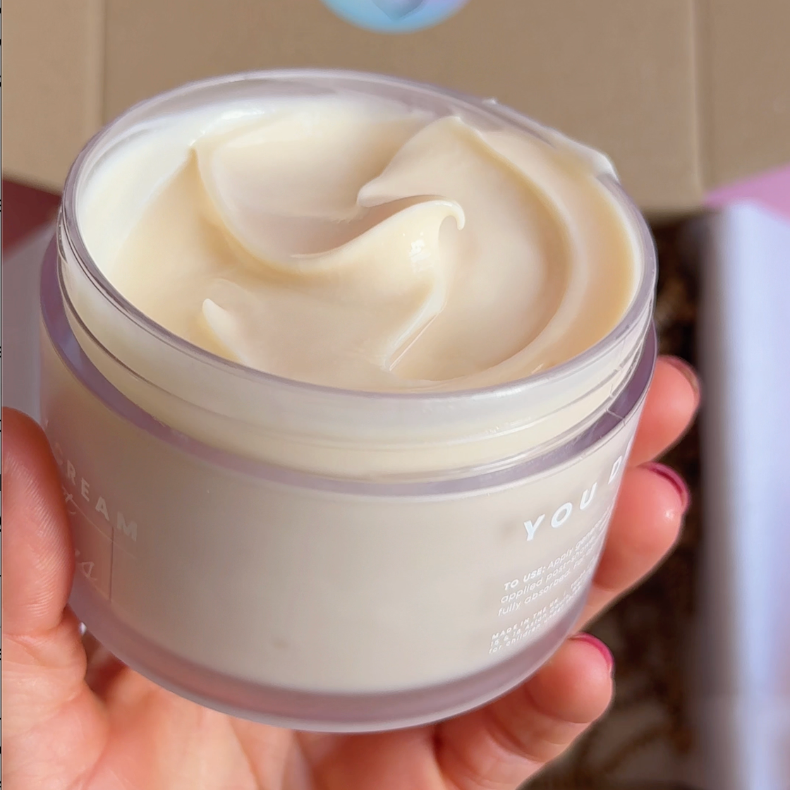A hand holding a clear jar filled with a thick, creamy off-white cosmetic cream, with a kraft paper box and a pastel birthday sticker in the background.